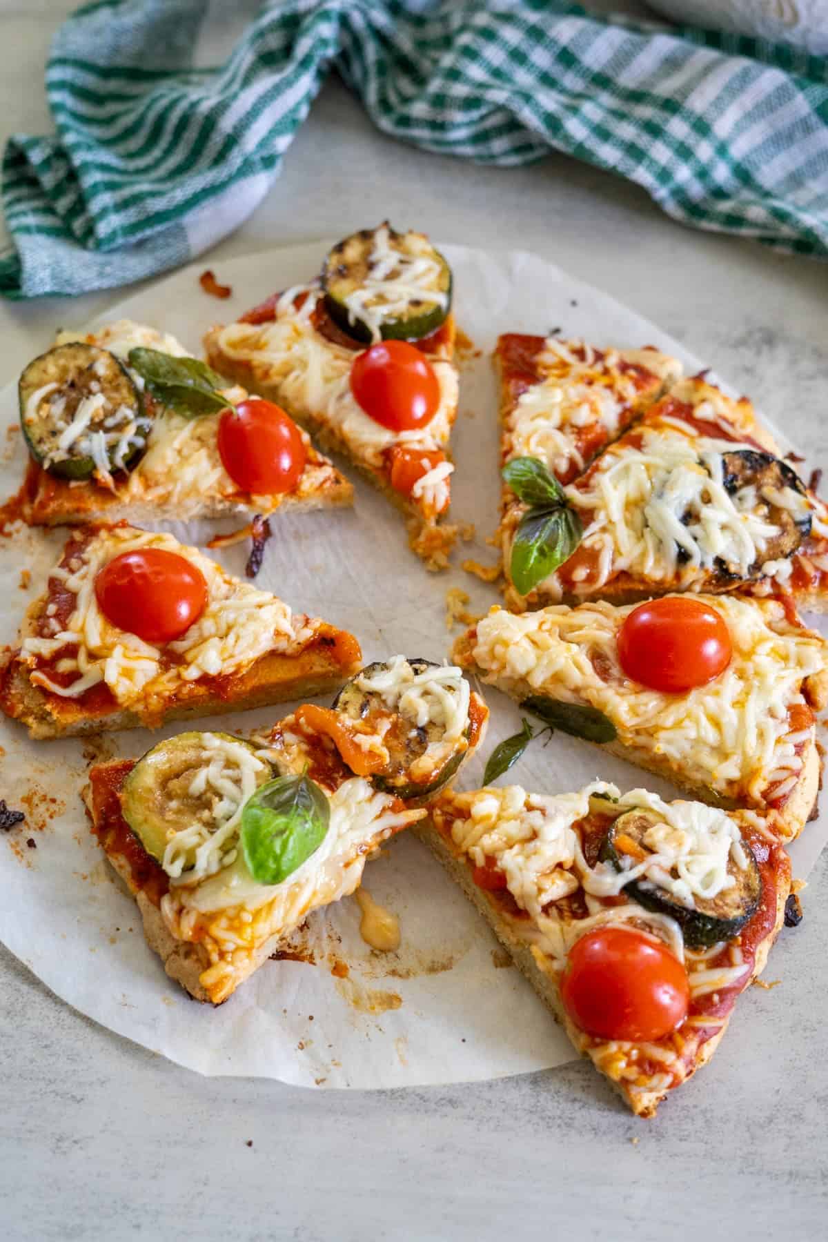 A sliced pizza made with almond flour, topped with cherry tomatoes, zucchini, basil leaves, and melted cheese, sits on parchment paper with a checkered cloth nearby.