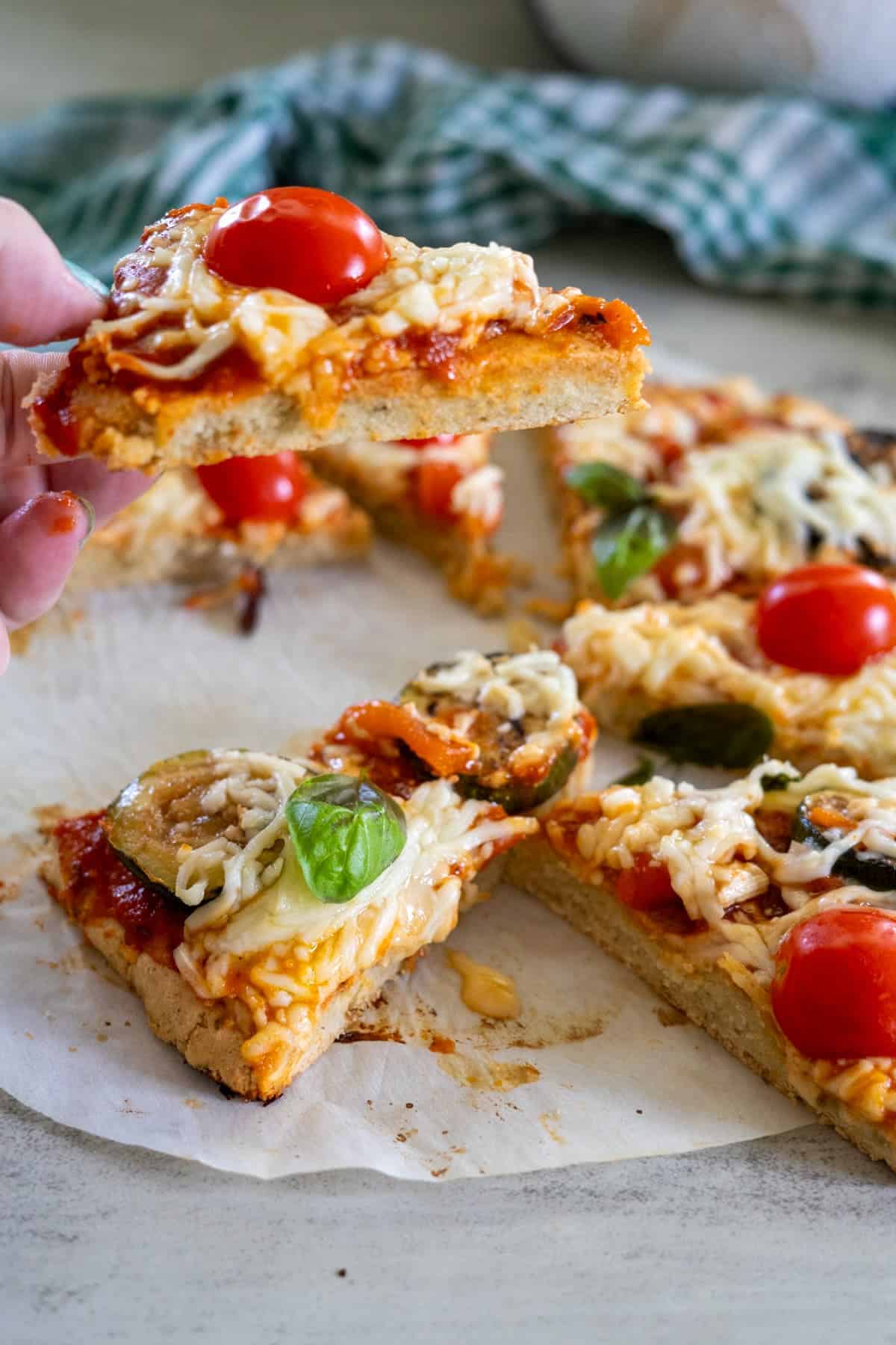 A hand holding a slice of pizza with toppings like cherry tomatoes, cheese, and basil showcases a crust made with almond flour. The rest of the pizza rests on parchment paper.