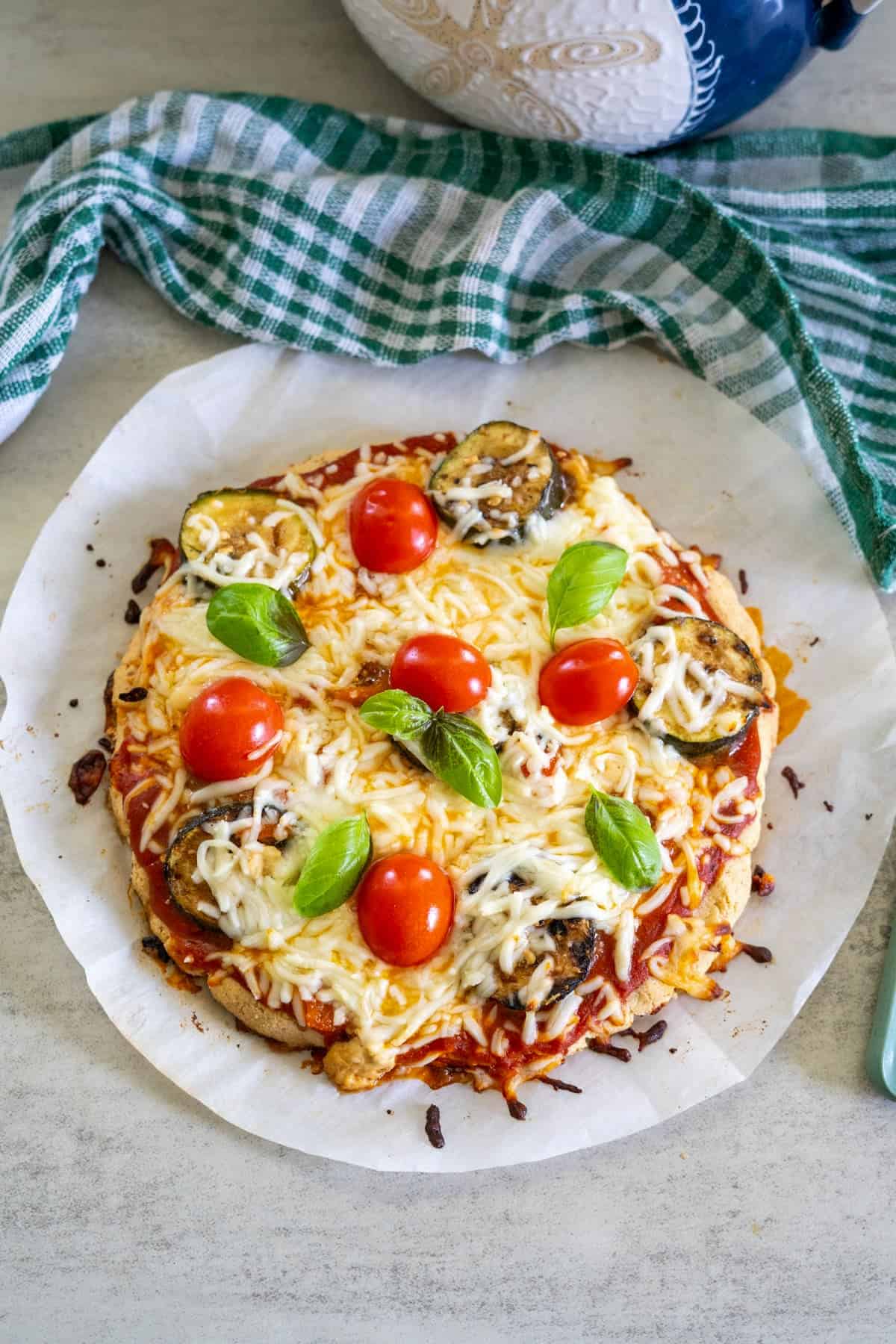 A homemade pizza with a crisp almond flour crust, topped with melted cheese, cherry tomatoes, eggplant slices, and fresh basil leaves on parchment paper, next to a green and white checkered cloth.