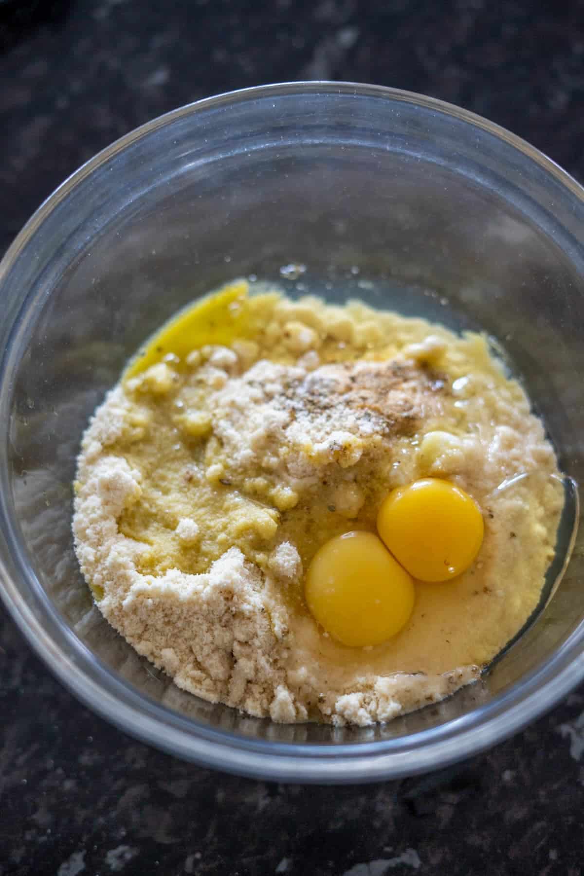 A glass bowl with almond flour, sugar, eggs, and mashed bananas rests on a dark surface.