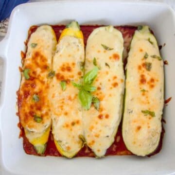 Three baked zucchini halves topped with melted cheese and basil in a white baking dish on tomato sauce.