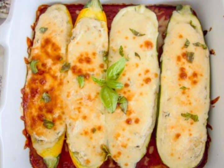 Three baked zucchini halves topped with melted cheese and basil in a white baking dish on tomato sauce.