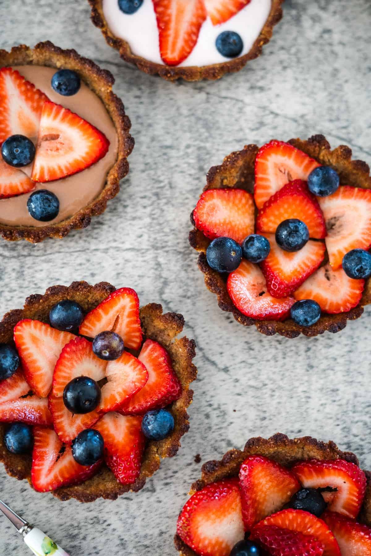 Five small keto berry tarts topped with sliced strawberries and blueberries, arranged on a speckled gray surface. Two tarts have a brown filling, and one has a white filling.