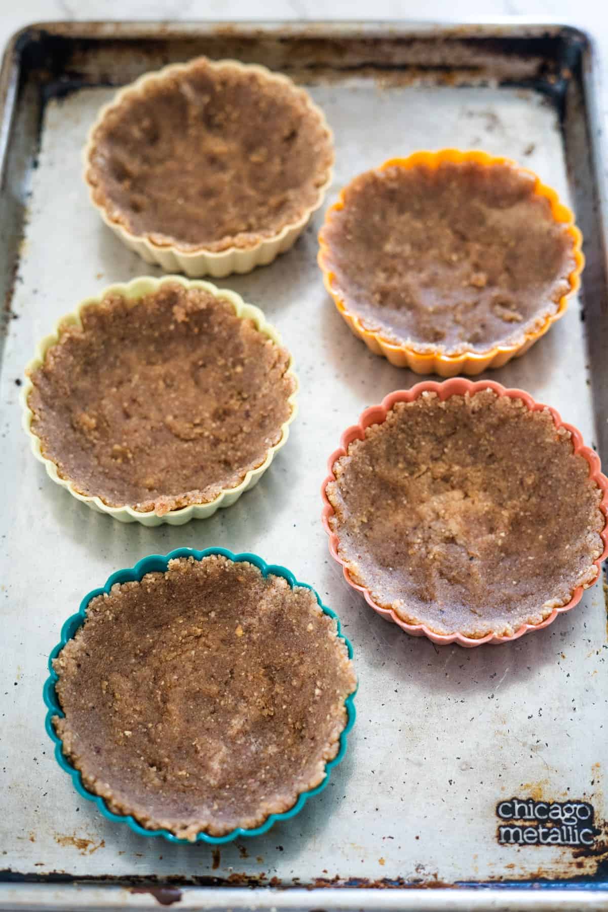 Five unbaked tart crusts in colorful silicone molds are placed on a metal baking sheet, ready to become delicious keto berry tarts. The baking sheet appears to be slightly worn.