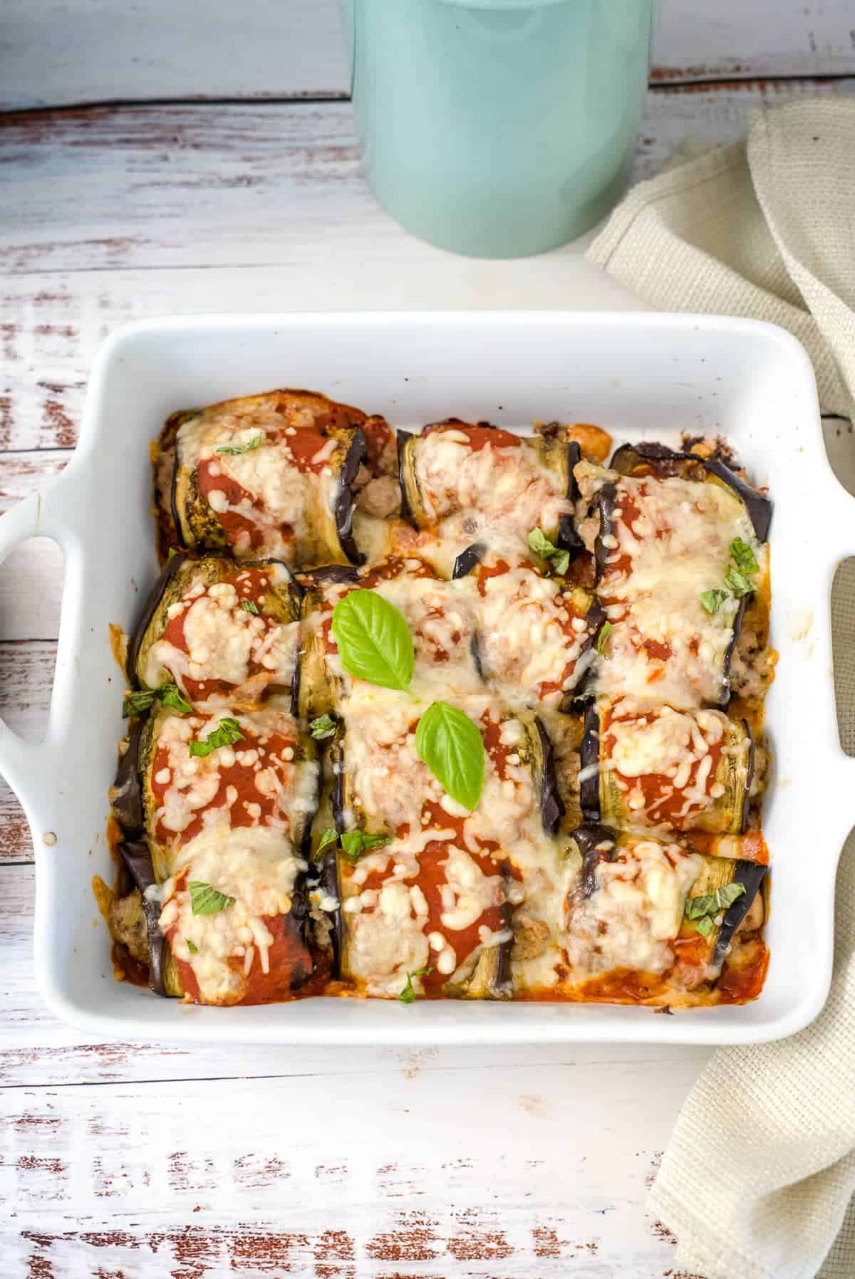 A white baking dish with nine portions of baked eggplant rollatini, some filled with ground turkey, topped with tomato sauce, melted cheese, and fresh basil leaves on a rustic wooden table with a white cloth.