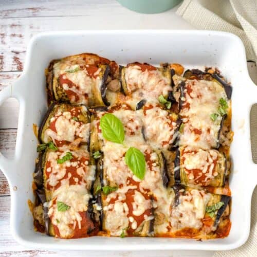 A white baking dish containing stuffed eggplant rolls with ground turkey, topped with melted cheese and garnished with fresh basil leaves.