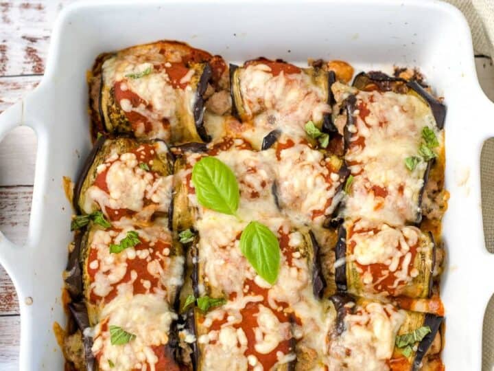 A white baking dish containing stuffed eggplant rolls with ground turkey, topped with melted cheese and garnished with fresh basil leaves.