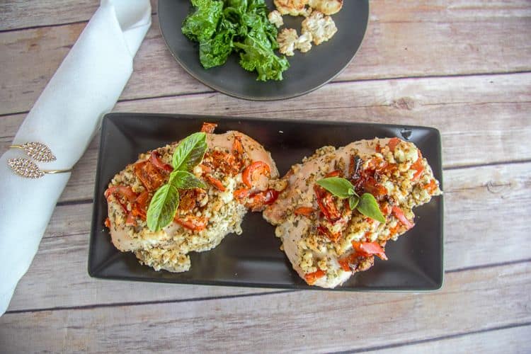 Two baked feta chicken breast fillets topped with a tomato-herb mixture and garnished with basil leaves on a rectangular black plate. Beside it is a plate of greens and a rolled napkin with a silver napkin ring.