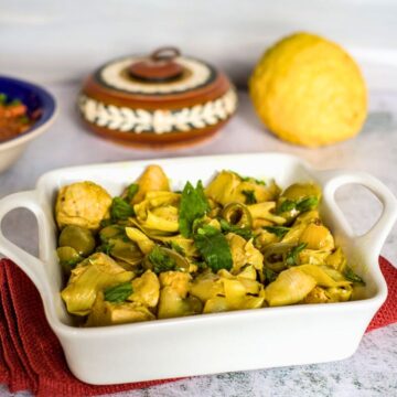 A white ceramic dish contains a cooked Keto Moroccan chicken meal with cubed chicken, artichokes, olives, and herbs. In the background, there is a round bread and a painted container.