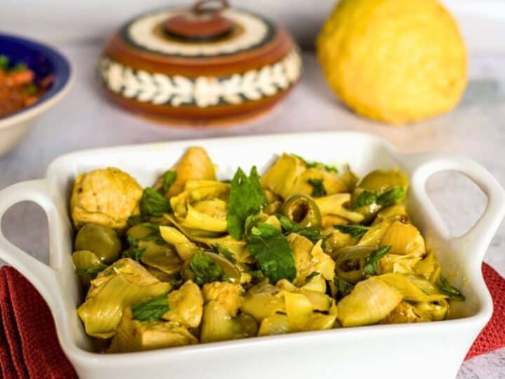 A white ceramic dish contains a cooked Keto Moroccan chicken meal with cubed chicken, artichokes, olives, and herbs. In the background, there is a round bread and a painted container.