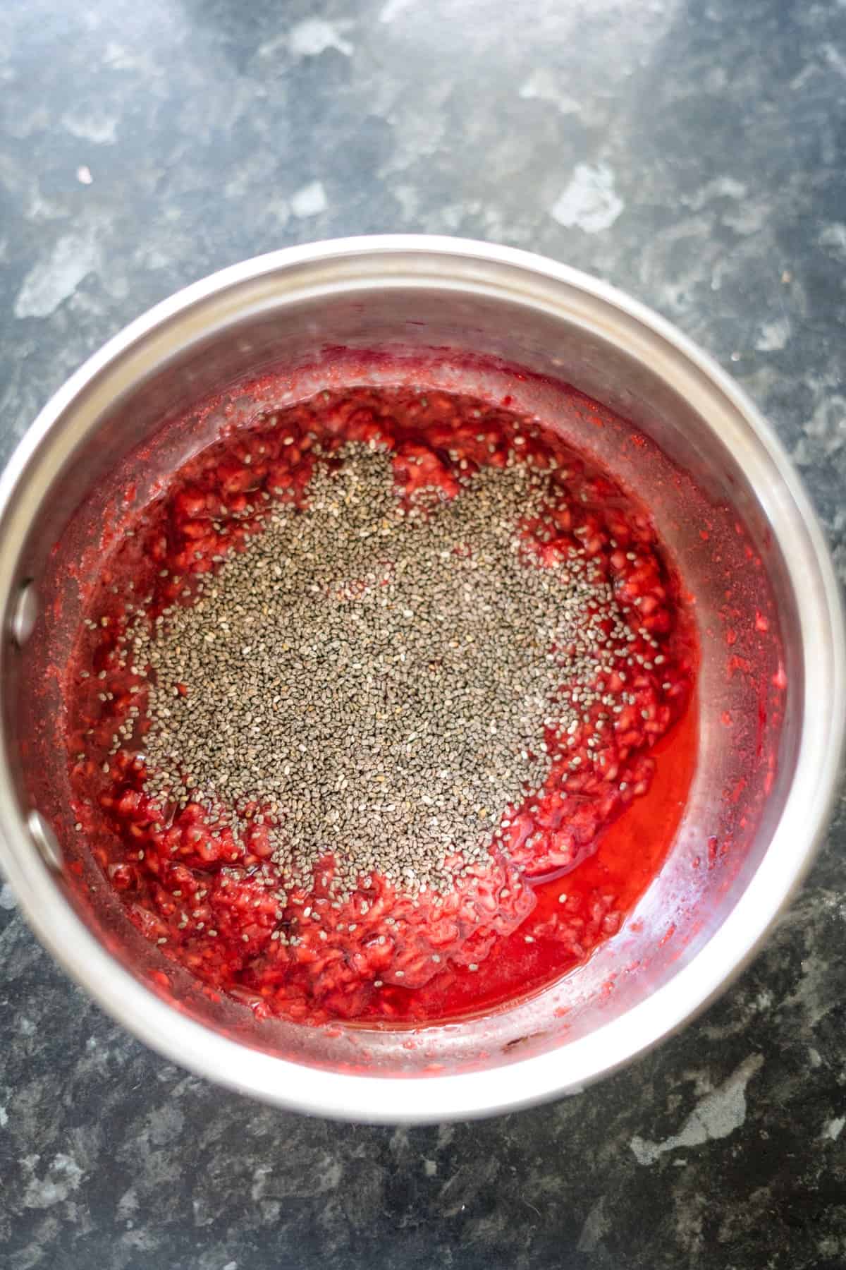 A stainless steel pot contains a mixture of mashed raspberries and chia seeds, creating a delicious raspberry chia jam on the black marble countertop.