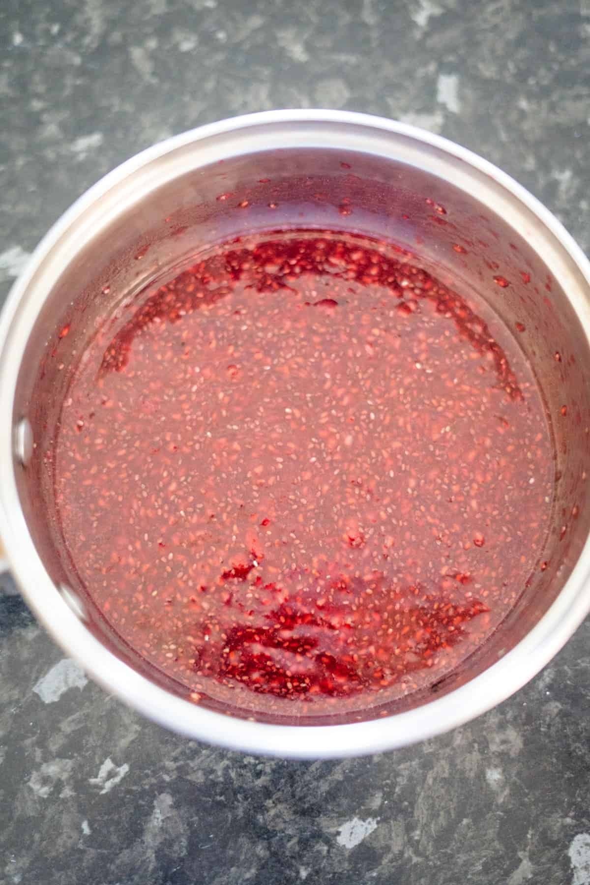 A pot filled with red raspberry chia jam mixture sits on a dark, marbled countertop.