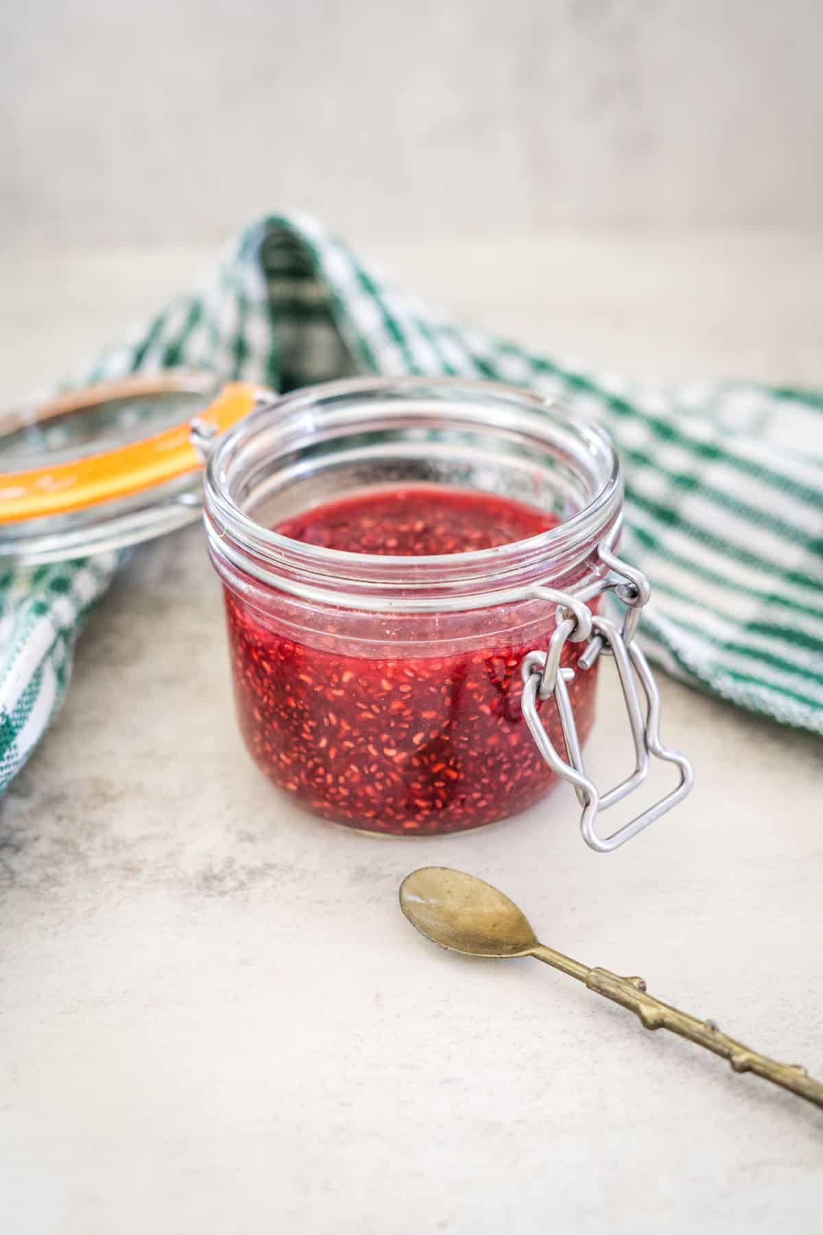 A glass jar filled with raspberry chia jam sits on a light surface near a closed lid and a green-striped towel. A small spoon with a thin handle lies in front of the jar.