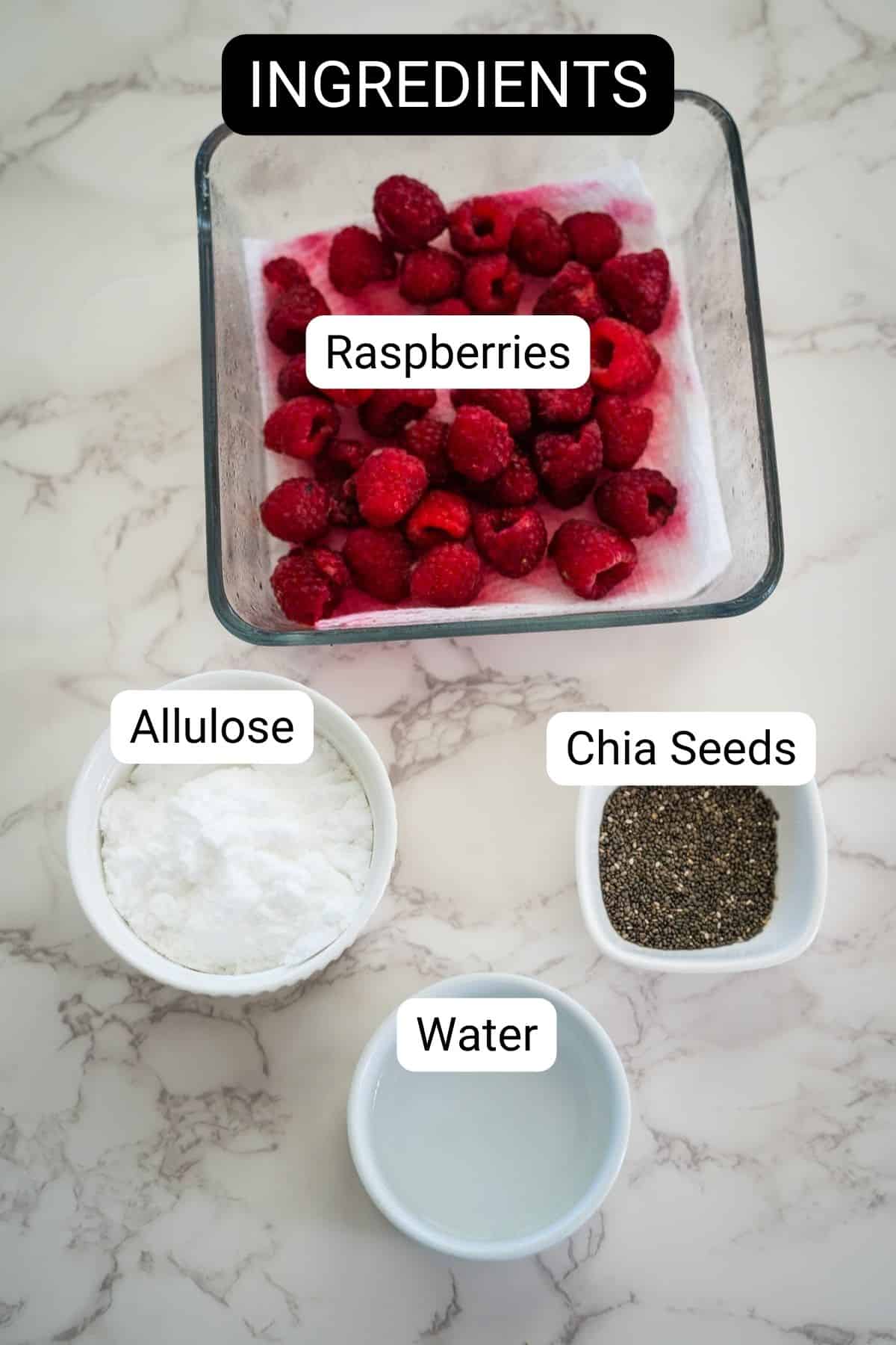 Ingredients for a raspberry chia jam recipe displayed on a marble surface: a bowl of raspberries, a bowl of allulose, a bowl of chia seeds, and a bowl of water.