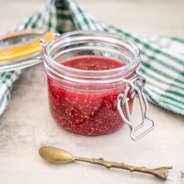 A glass jar filled with raspberry chia jam, placed on a light-colored surface next to a small decorative spoon and a green and white striped cloth.