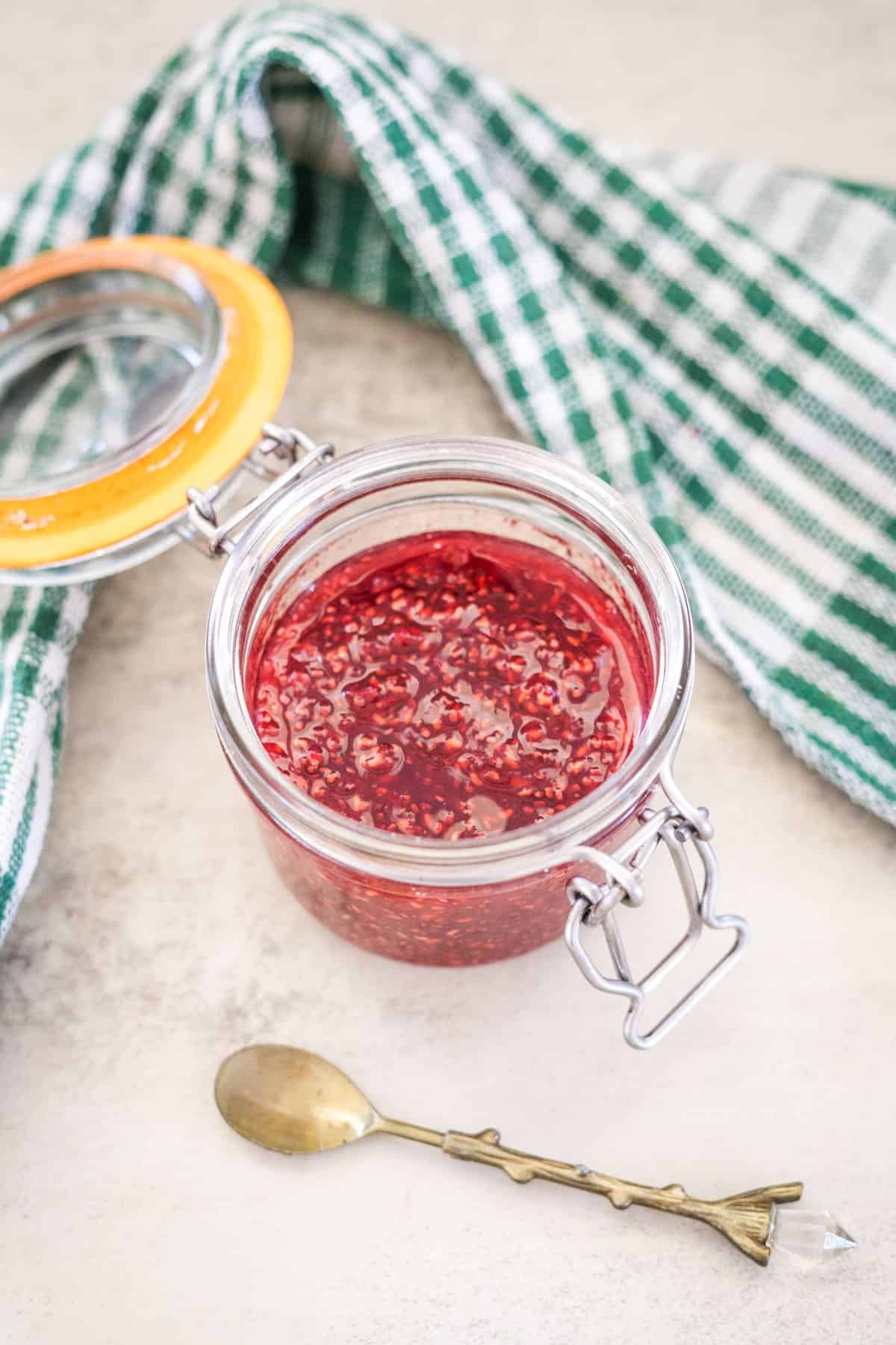 An open glass jar of raspberry chia jam sits next to a green and white checkered cloth, accompanied by a small ornate spoon on a light surface.