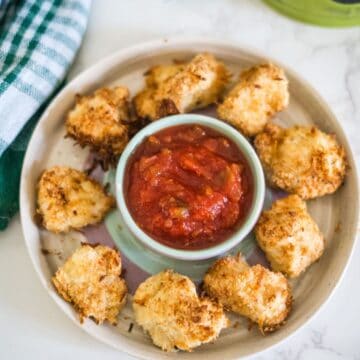 A plate of oven-baked cauliflower bites arranged around a small bowl of marinara sauce, with a green and white checkered cloth placed beside the plate.