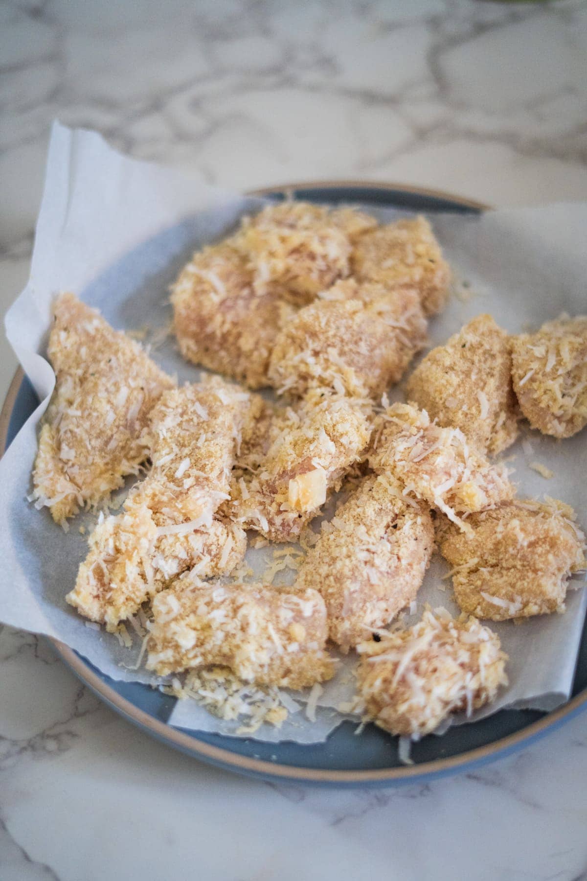 A plate with pieces of breaded, uncooked chicken on parchment paper, placed on a marble countertop.