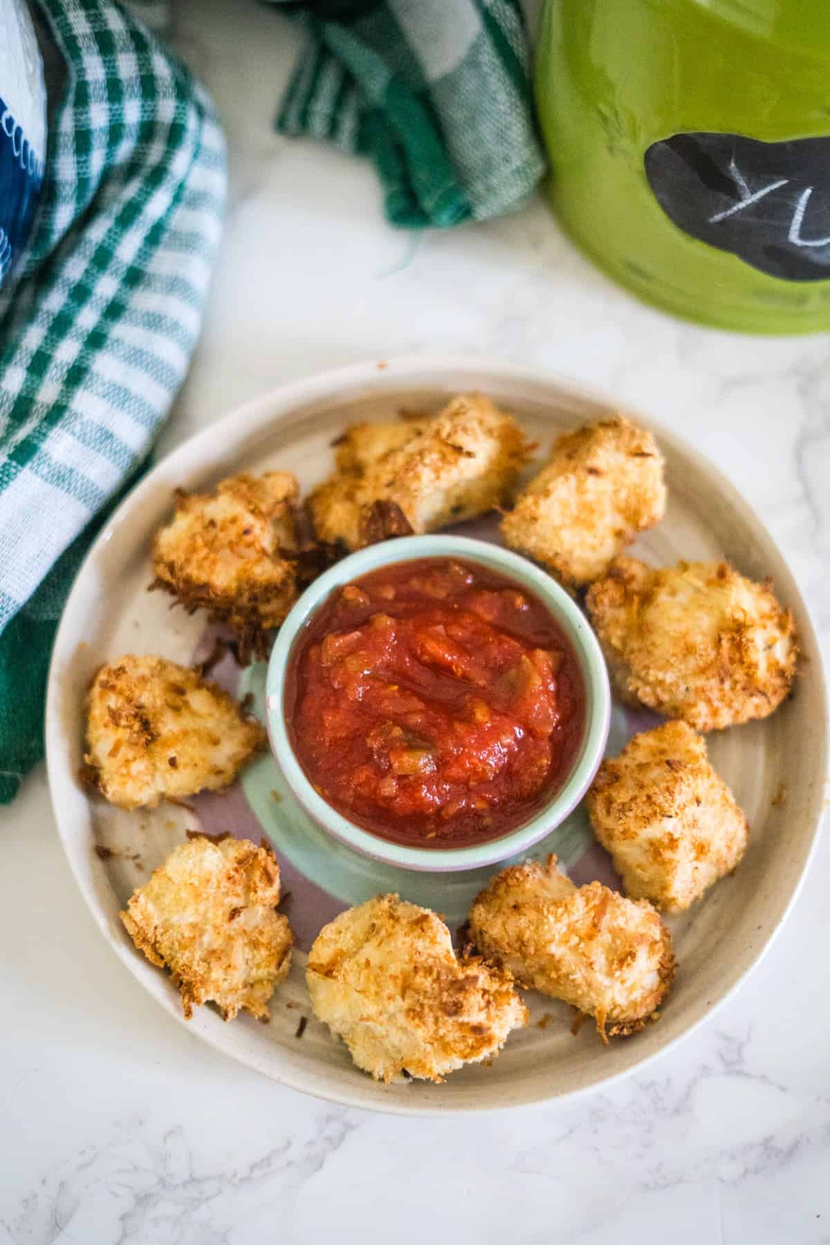 A round plate with crispy, fried cauliflower bites surrounding a small bowl of red dipping sauce. A green and white cloth is partially visible in the background.