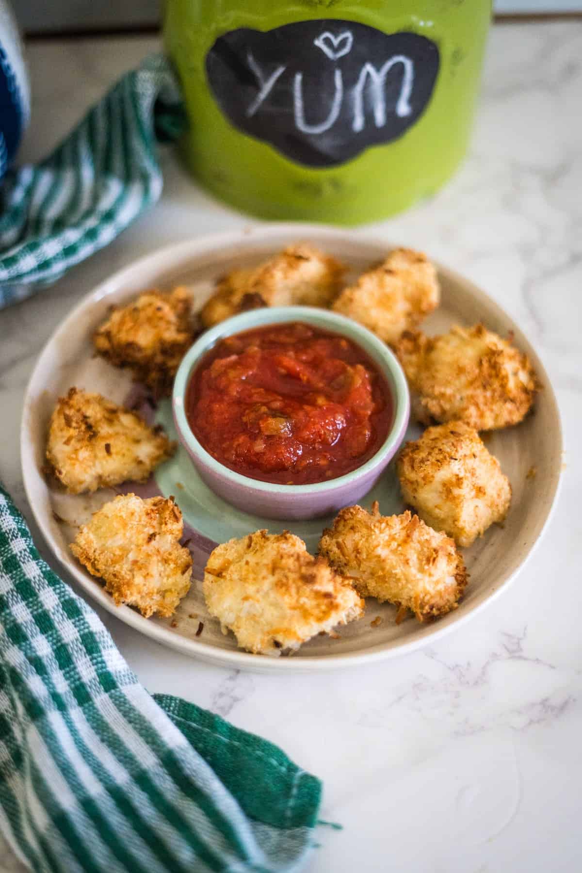 A plate of baked coconut shrimp surrounds a bowl of red dipping sauce, placed on a marble surface with a green-striped cloth nearby and a green jar labeled "YUM" in the background.