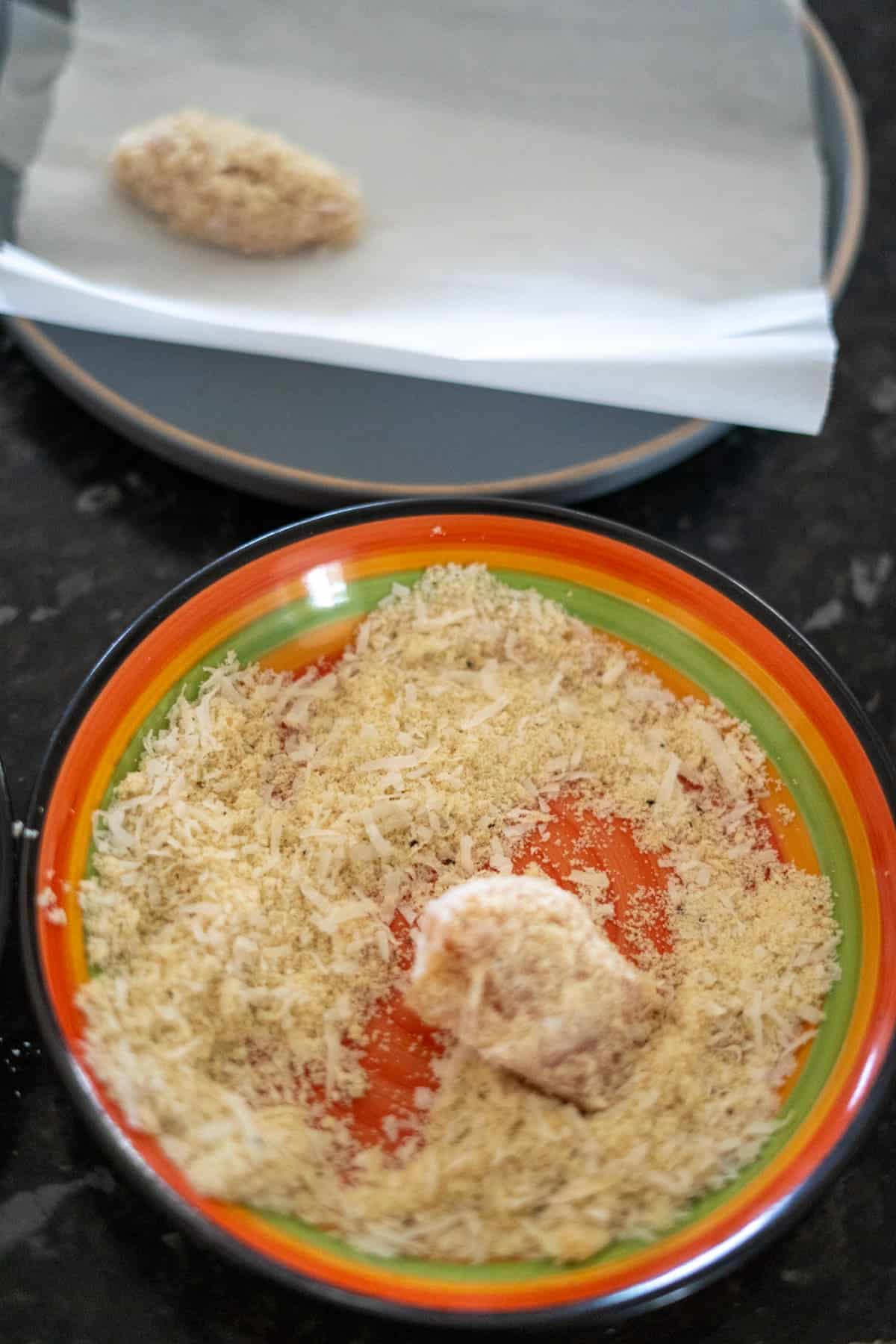 A bowl containing a breaded item partially covered in breadcrumbs and a plate in the background with a tray lined with parchment paper holding another breaded item.