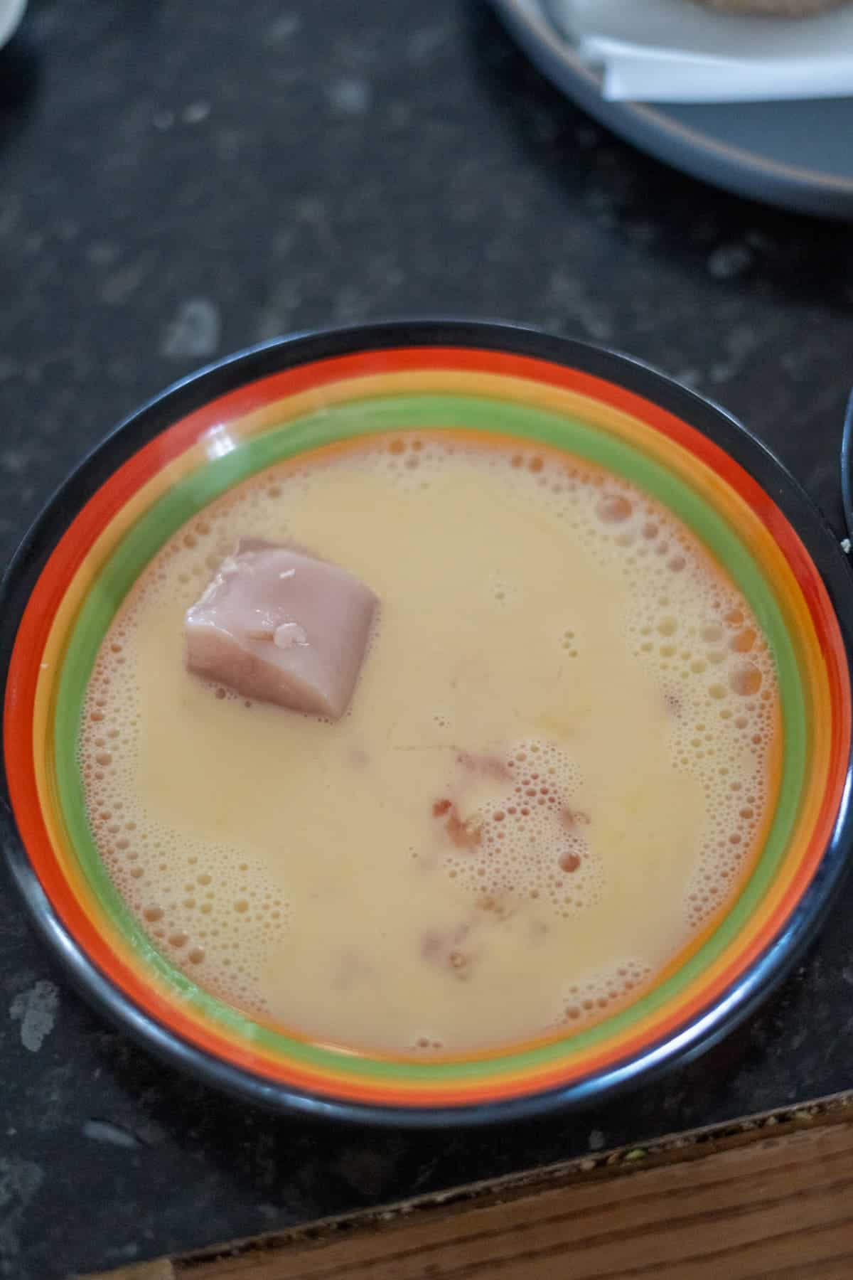 A bowl with a rainbow-striped rim containing a beige-colored liquid with bubbles and a small block of a solid substance.