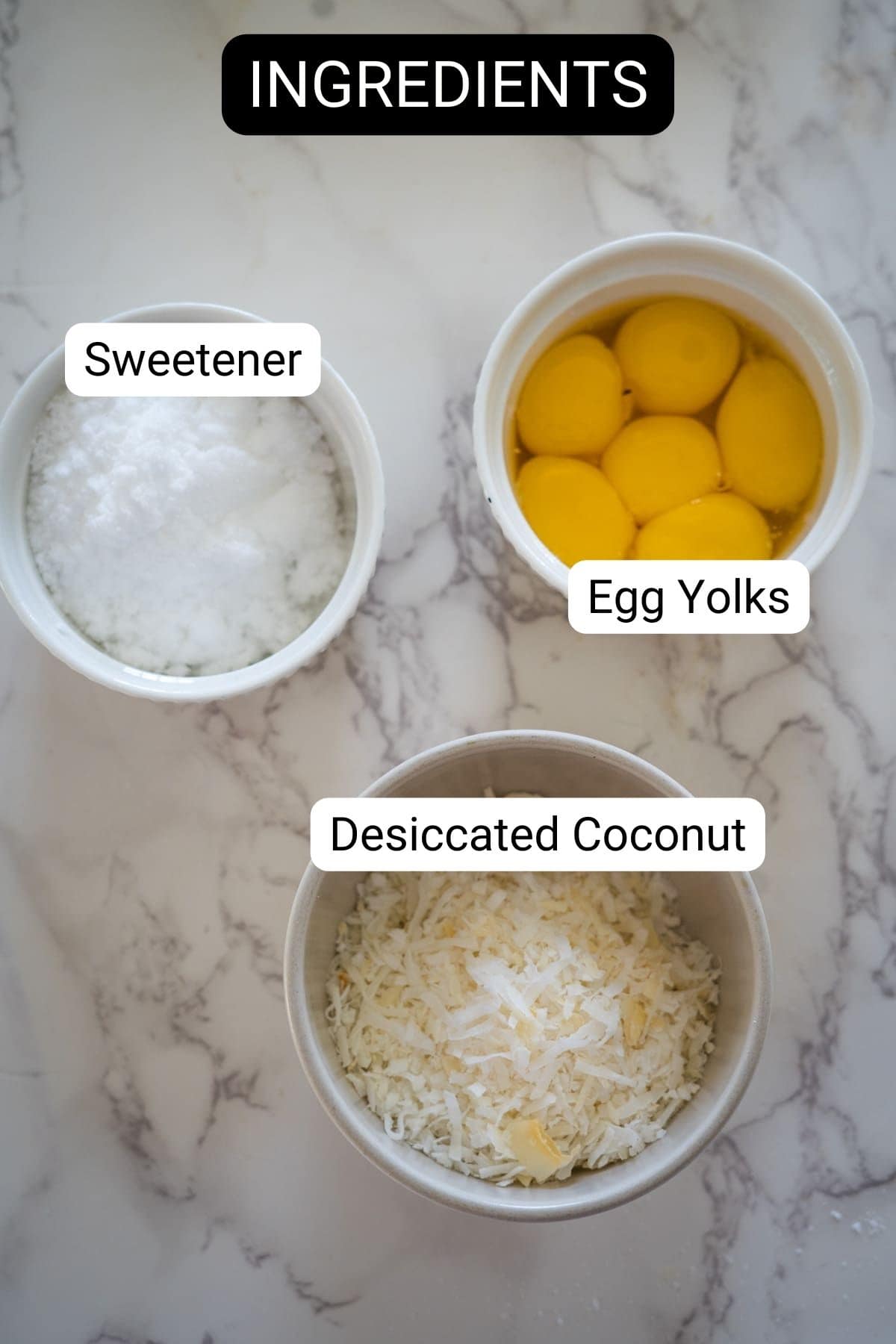 Top view of three bowls, each labeled with their contents: sweetener, egg yolks, and desiccated coconut—ingredients for a delicious keto coconut flan. The background is a white marble surface.