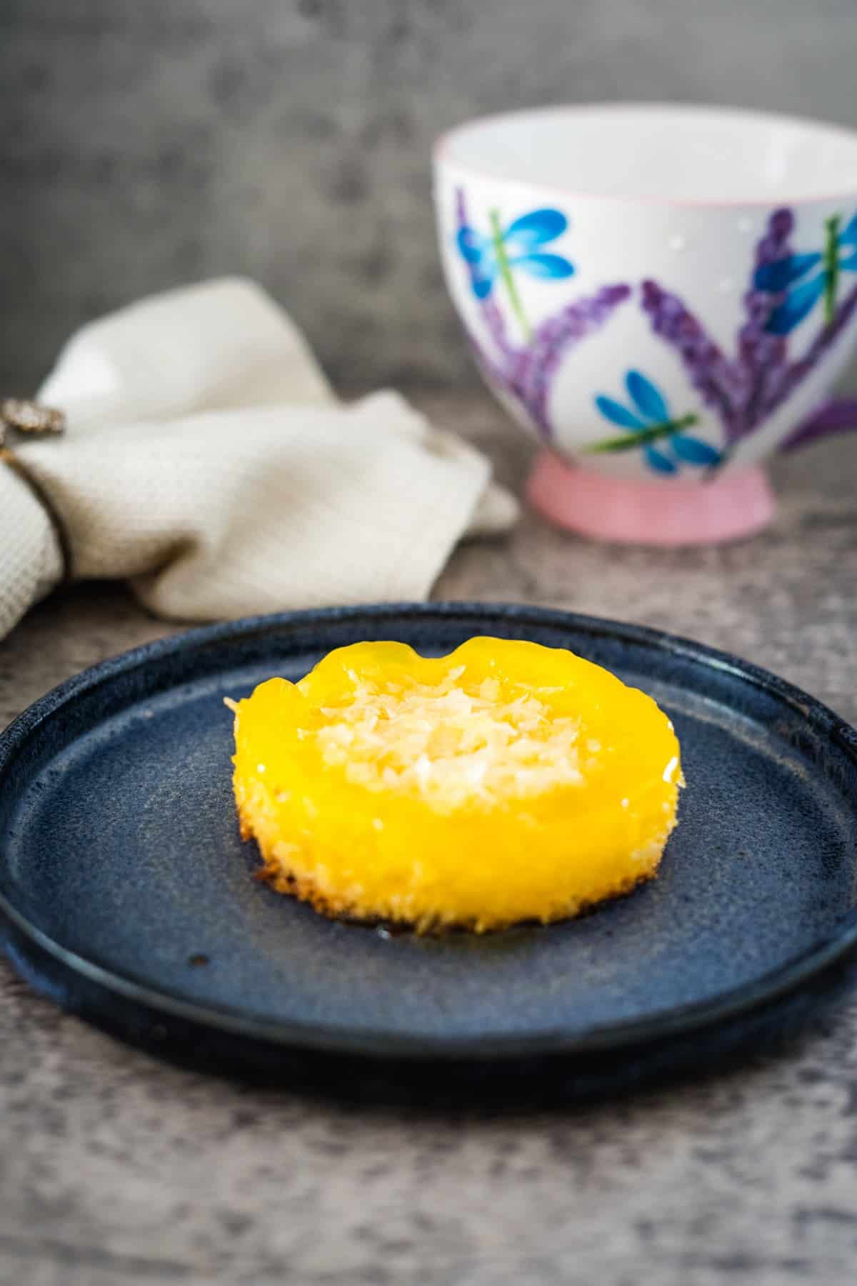 A round, yellow keto coconut flan topped with shredded coconut on a blue plate. A colorful mug and a beige cloth are in the background.