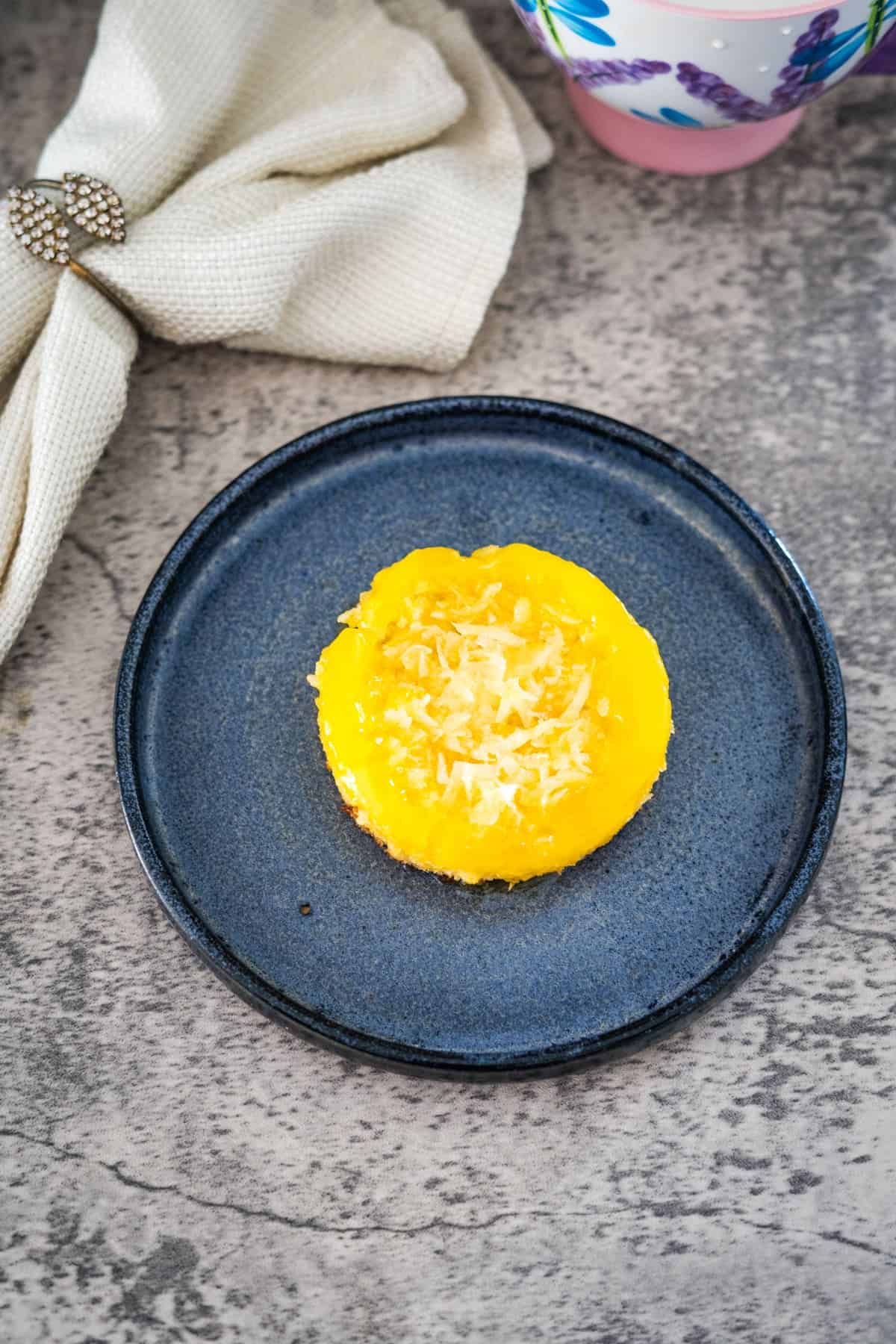 A single keto coconut flan topped with shredded cheese is placed on a dark blue plate with a beige napkin and a cup partially visible in the background.