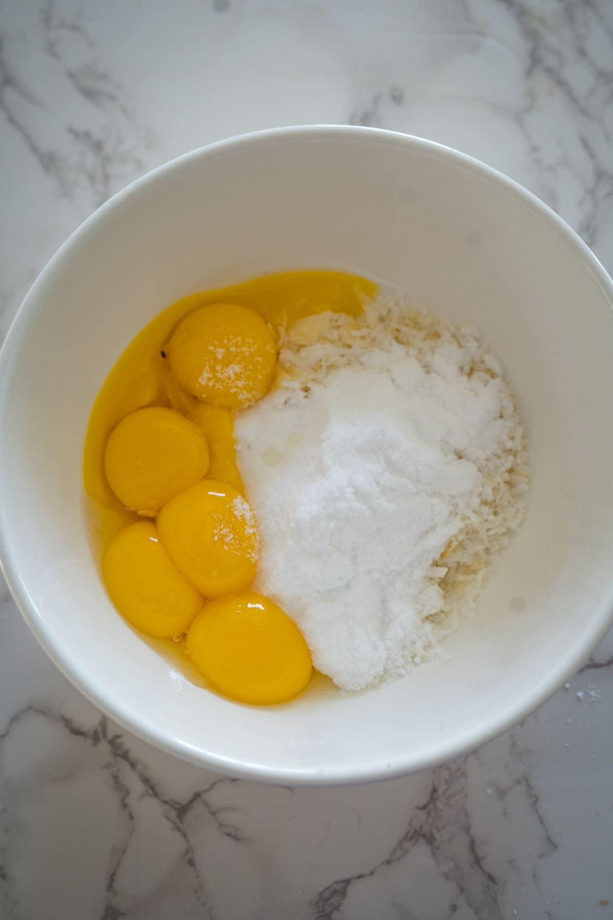 A white bowl containing egg yolks, granulated sugar, and shredded coconut—perfect ingredients for a keto coconut flan—rests on a marble countertop.