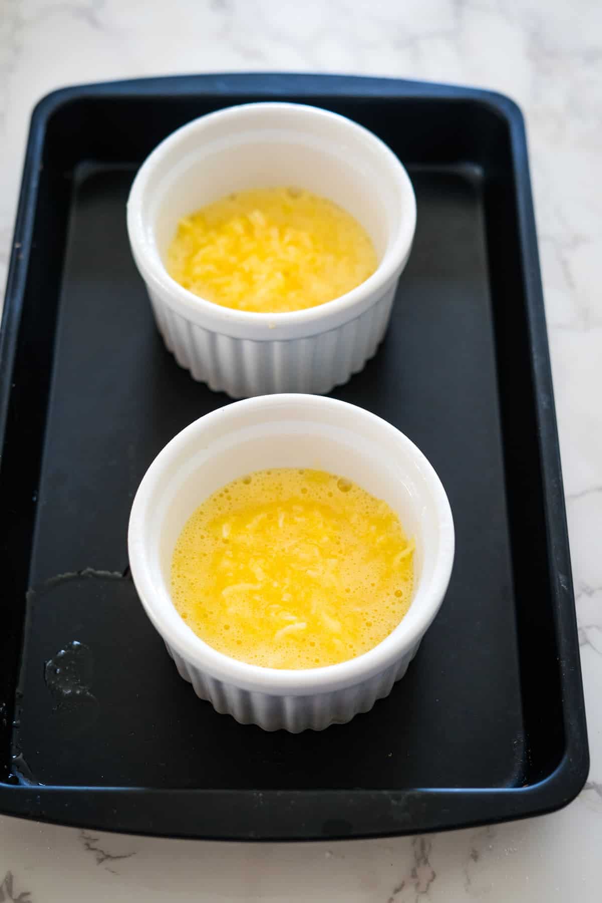 Two white ramekins filled with a yellow mixture of keto coconut flan sit on a black baking tray, placed on a white marble surface.