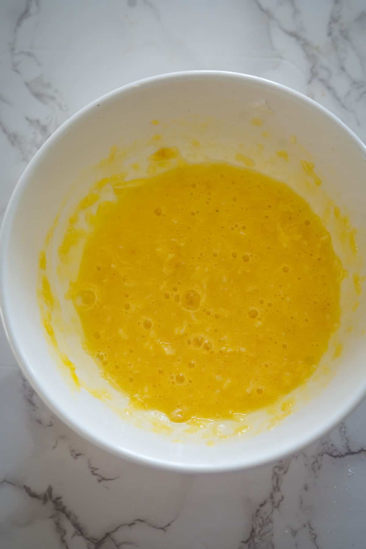 A white mixing bowl filled with a yellow, frothy liquid batter, perfect for making a keto coconut flan, sits on a marble surface.