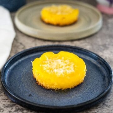 A round yellow keto coconut flan with a crumbly base and grated topping is served on a blue plate, with another similar dessert in the background on a light-colored plate.