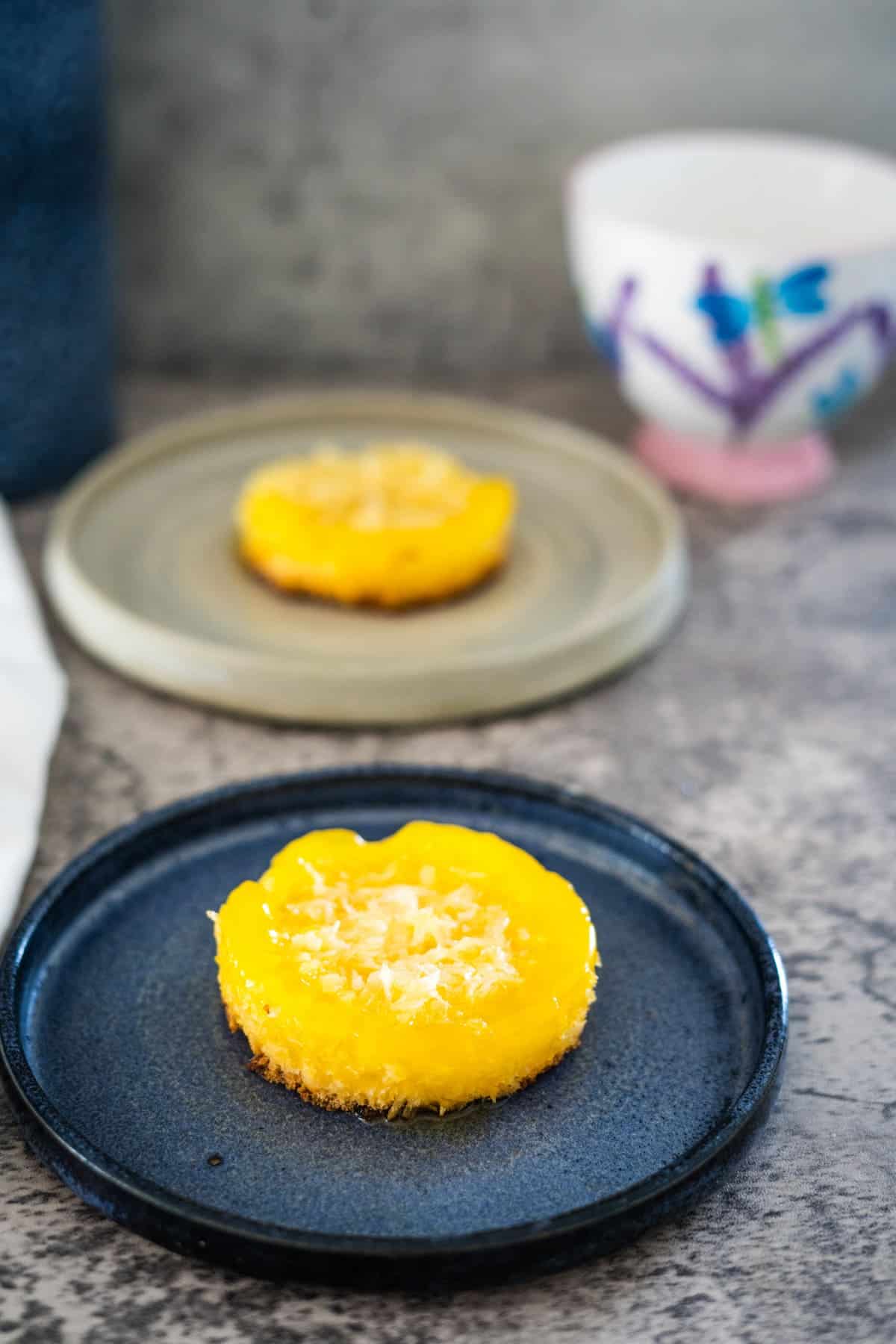 Two round yellow pastries are displayed on separate plates. The keto coconut flan in the foreground sits on a dark blue plate, while the one in the background rests on a light grey plate.