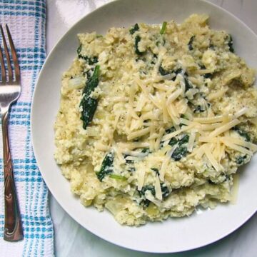 A bowl of creamy risotto made with cauliflower rice and spinach, topped with shredded parmesan cheese, beside a fork on a cloth napkin.