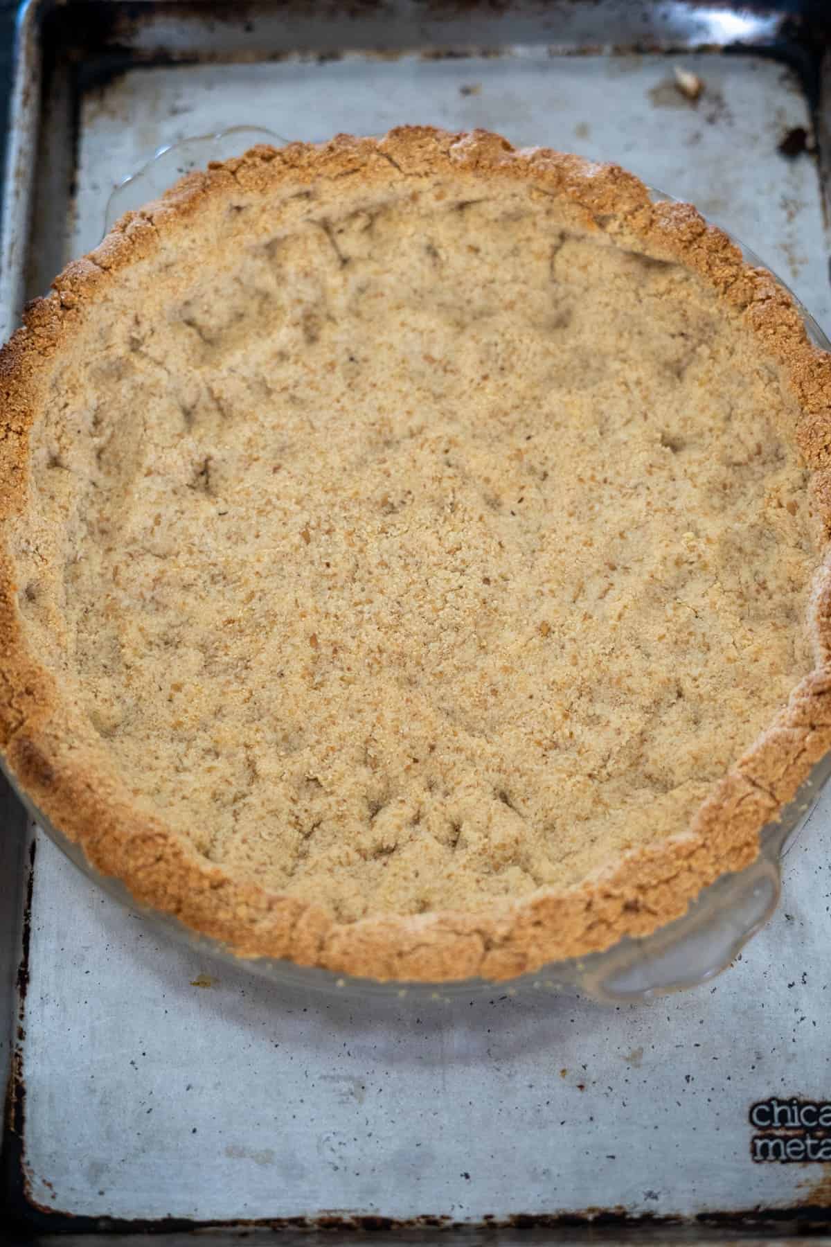 A baked pie crust in a glass pie dish sits on a metal baking sheet, ready to be filled with a rich broccoli and blue cheese quiche. The crust appears golden-brown and evenly cooked.