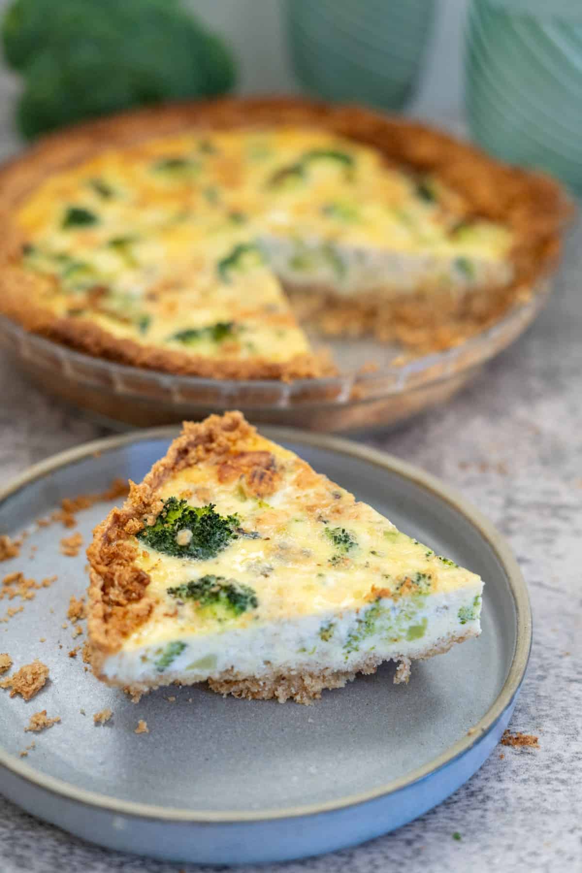 A slice of broccoli and blue cheese quiche on a grey plate with the remaining quiche in the background.