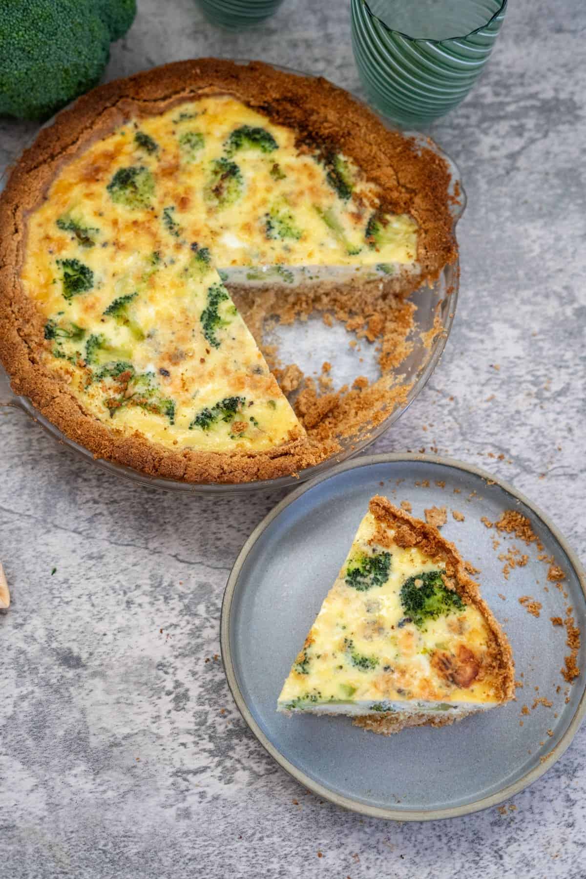 A broccoli and blue cheese quiche with one slice removed and placed on a gray plate. The pie is on a gray surface with a green glass in the background.