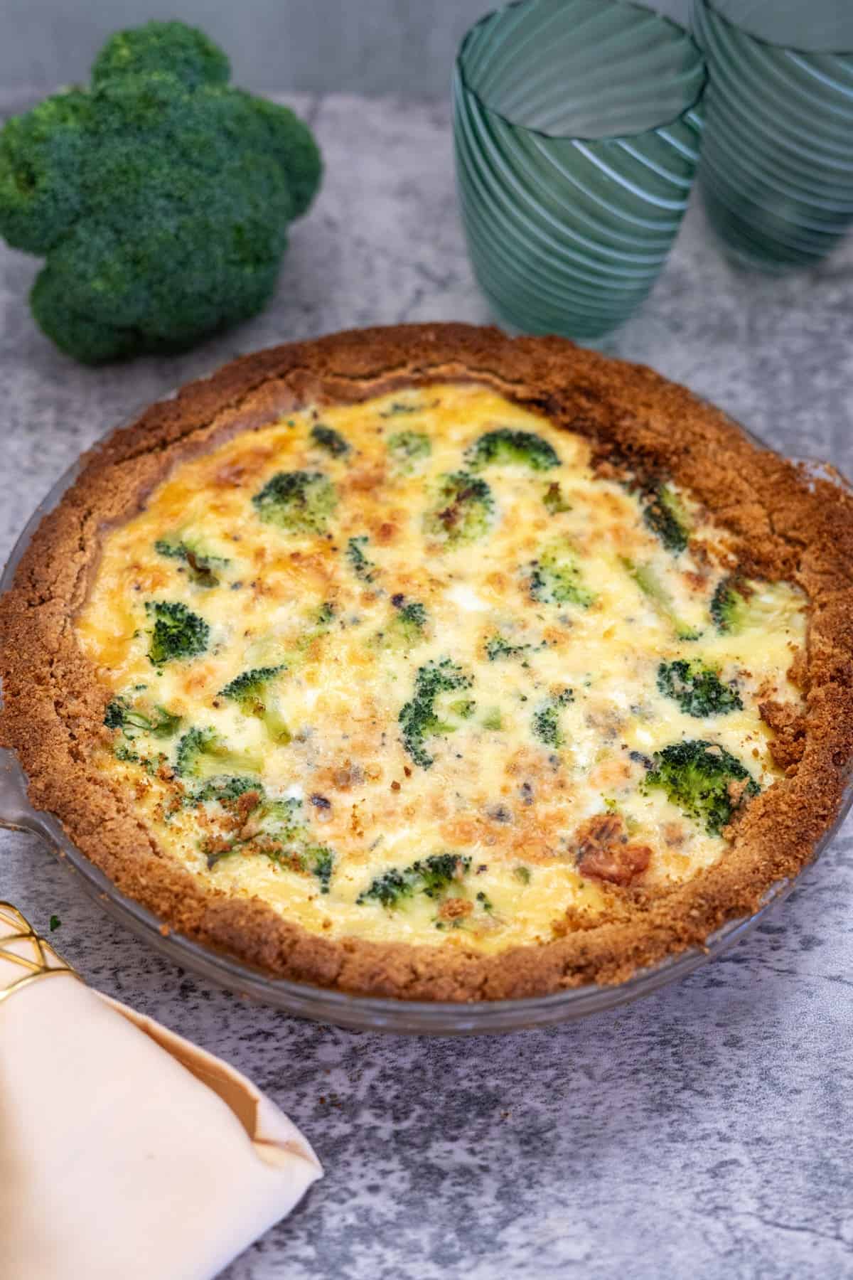 A broccoli and blue cheese quiche with a golden-brown crust sits on a gray surface next to a head of broccoli, two green glasses, and a folded napkin.