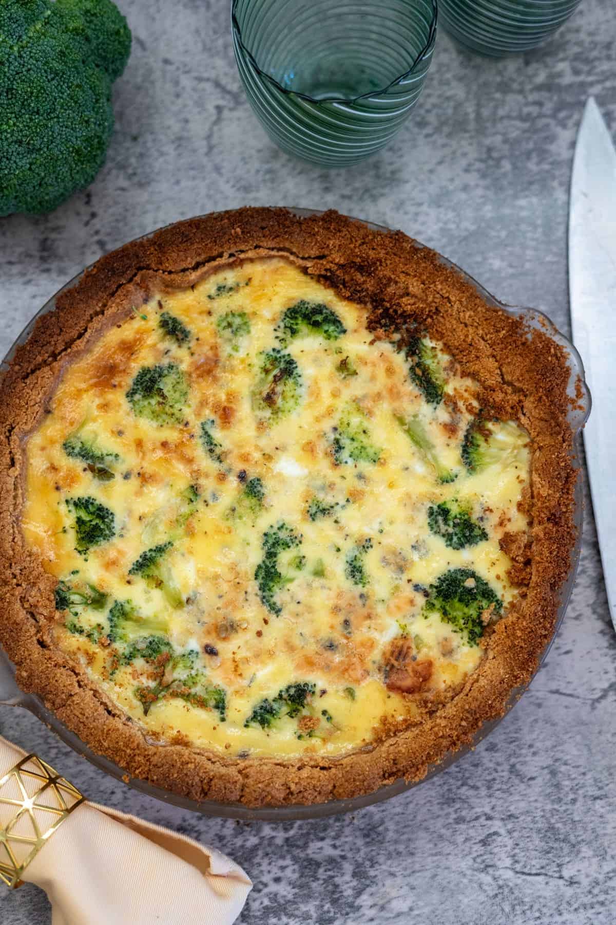 A broccoli and blue cheese quiche with a golden-brown crust, placed on a gray surface beside two green glasses and a knife.