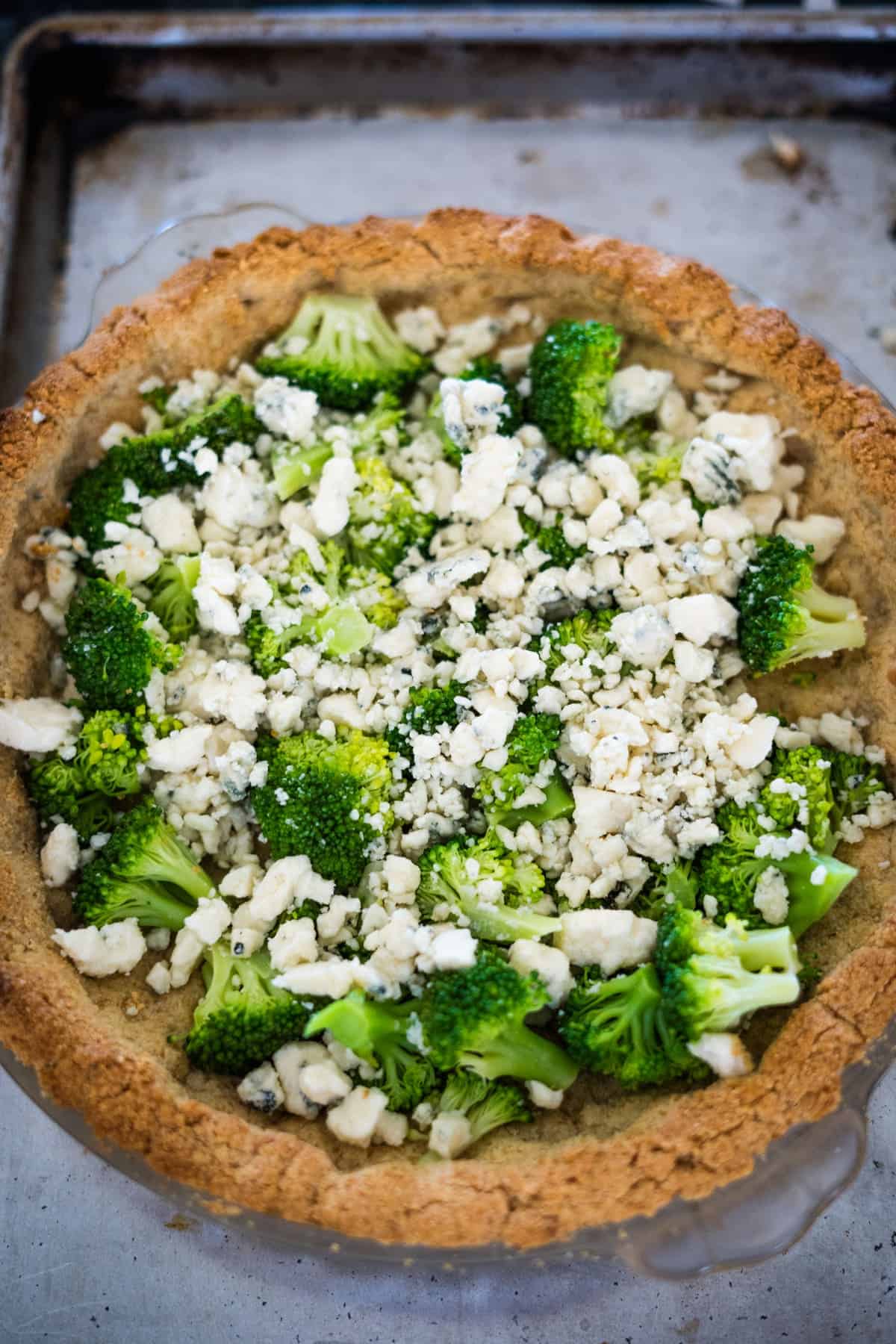 A broccoli and blue cheese quiche base filled with broccoli florets and crumbled cheese, ready to be baked, on a baking tray.