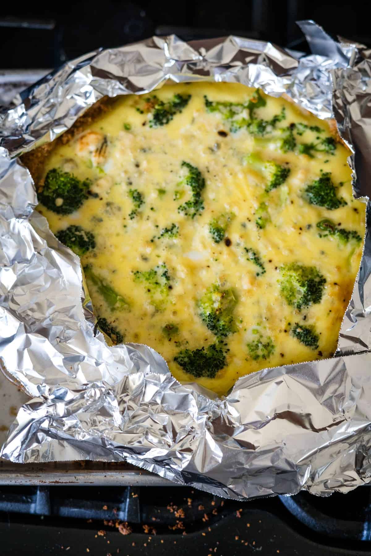 A baked broccoli and blue cheese quiche, partially wrapped in aluminum foil, rests on a stovetop.