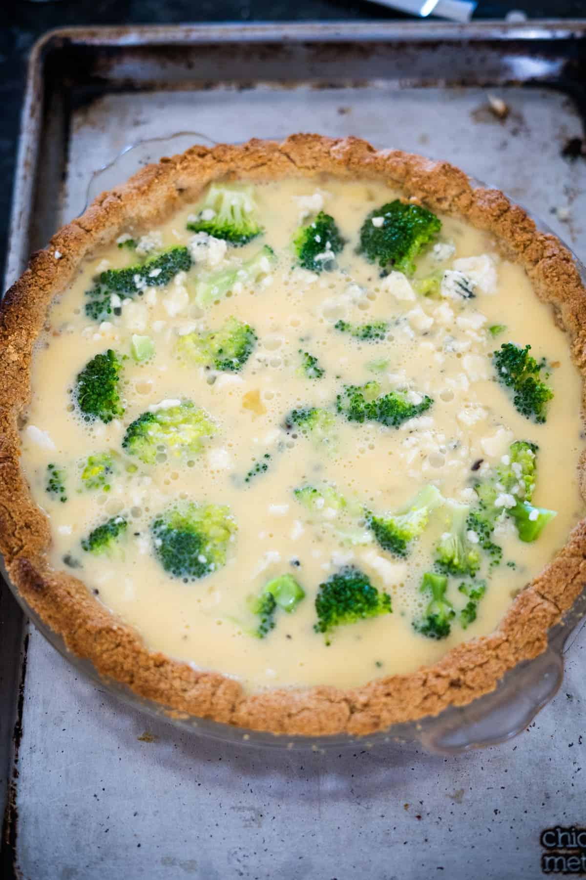 A broccoli and blue cheese quiche in a pie crust sits on a baking sheet, ready to be baked.