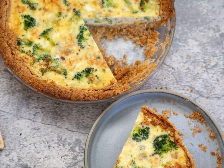 A broccoli and blue cheese quiche with a slice removed, placed on a separate plate next to it. The quiche filling includes broccoli and melted cheese, and the crust is golden brown.