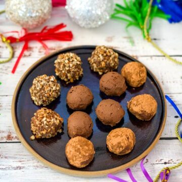 A black plate with twelve round pumpkin chocolate truffles, some coated in nuts and some in cocoa powder, on a festive table with colorful decorations in the background.