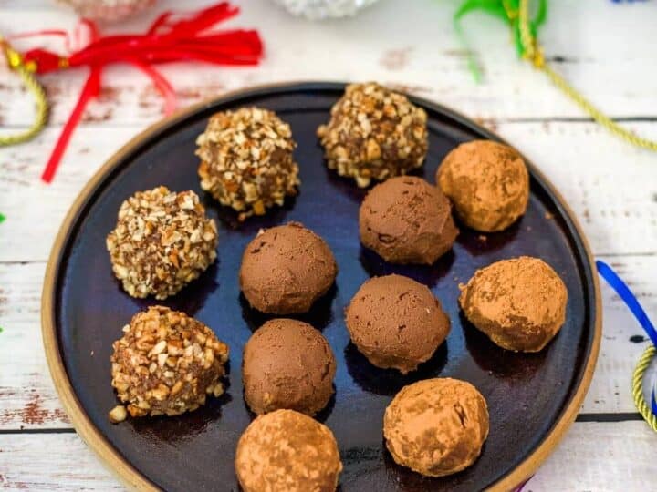 A black plate with twelve round pumpkin chocolate truffles, some coated in nuts and some in cocoa powder, on a festive table with colorful decorations in the background.