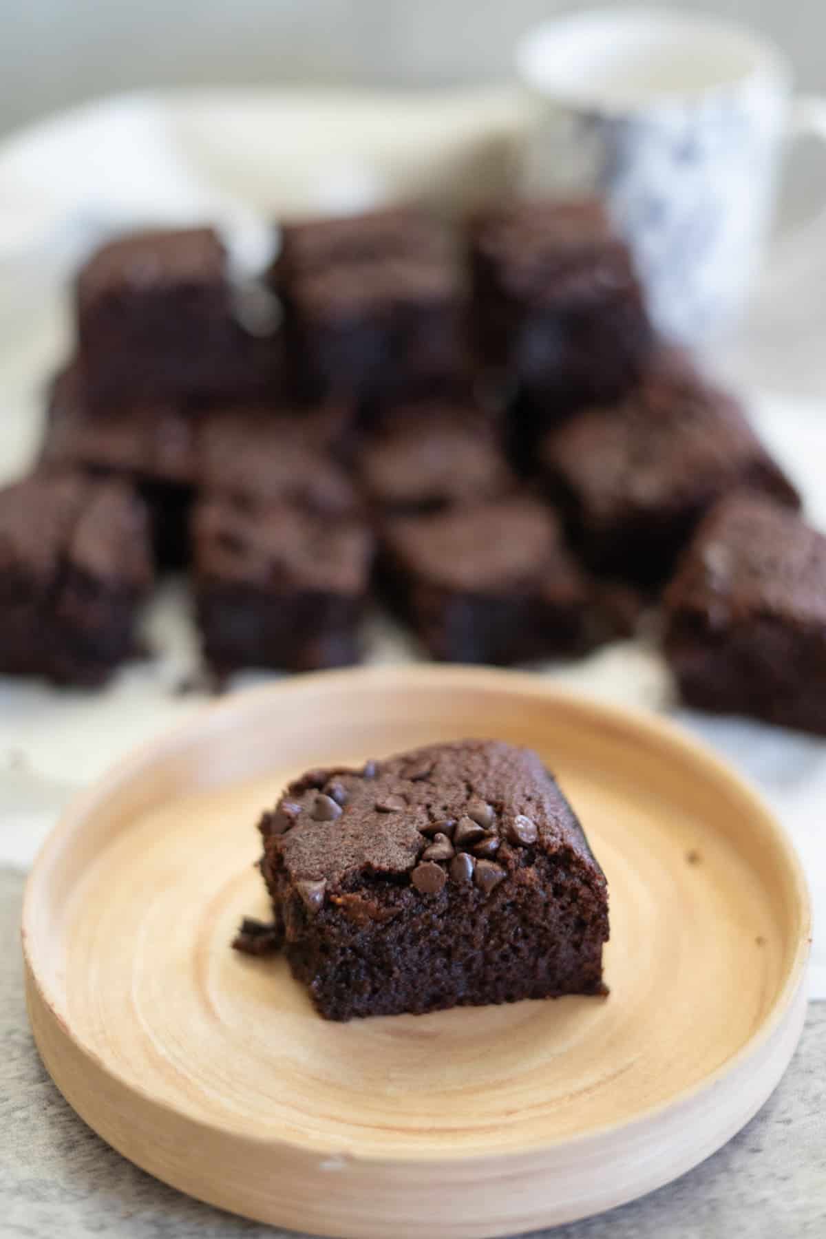 A wooden plate with a single chocolate brownie, in front of a blurred stack of brownies and a mug.