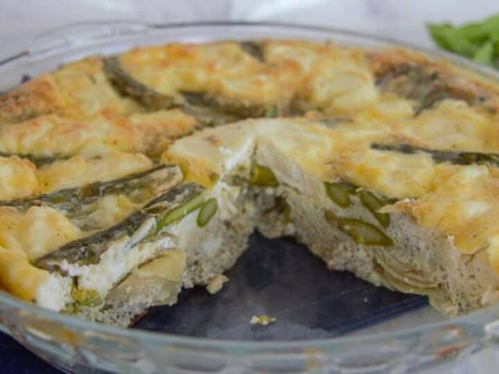 A glass pie dish showcasing a partially eaten crustless quiche, with visible green asparagus nestled in a golden-brown filling.