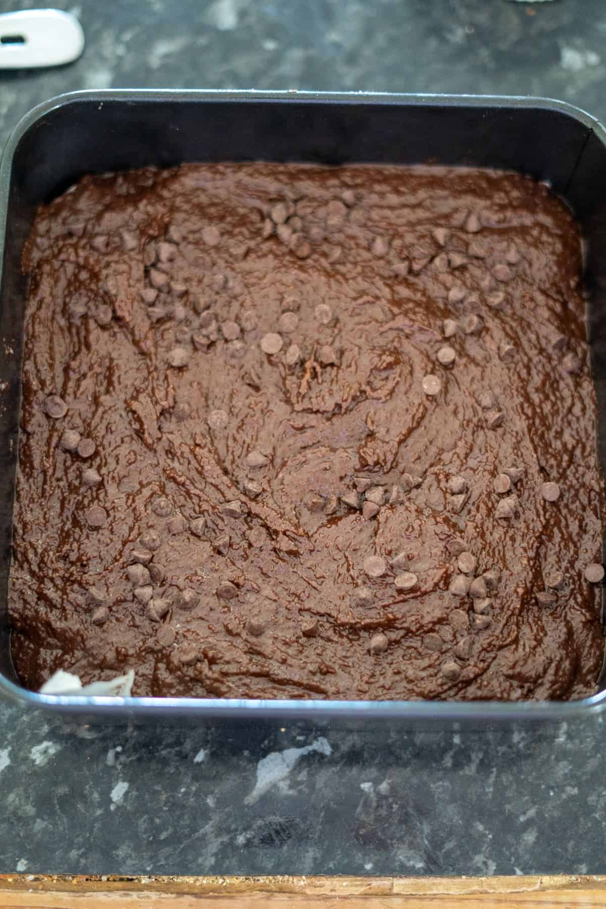 Brownie batter with chocolate chips spread in a square baking pan on a dark countertop.