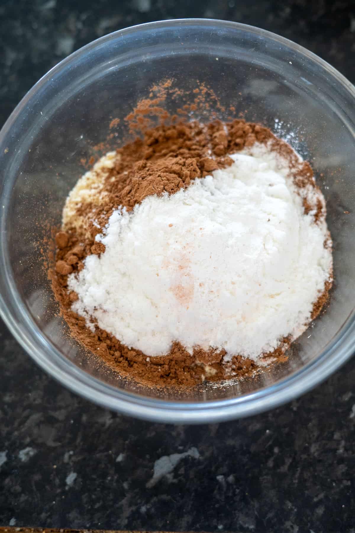 A glass bowl containing flour, cocoa powder, and sugar, set on a dark countertop.